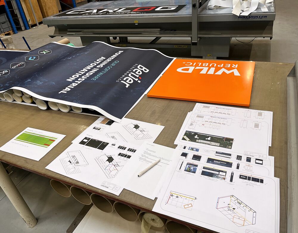 Standtegninger, grafik og færdige skilteelementer samlet på et arbejdsbord hos Wieben Design